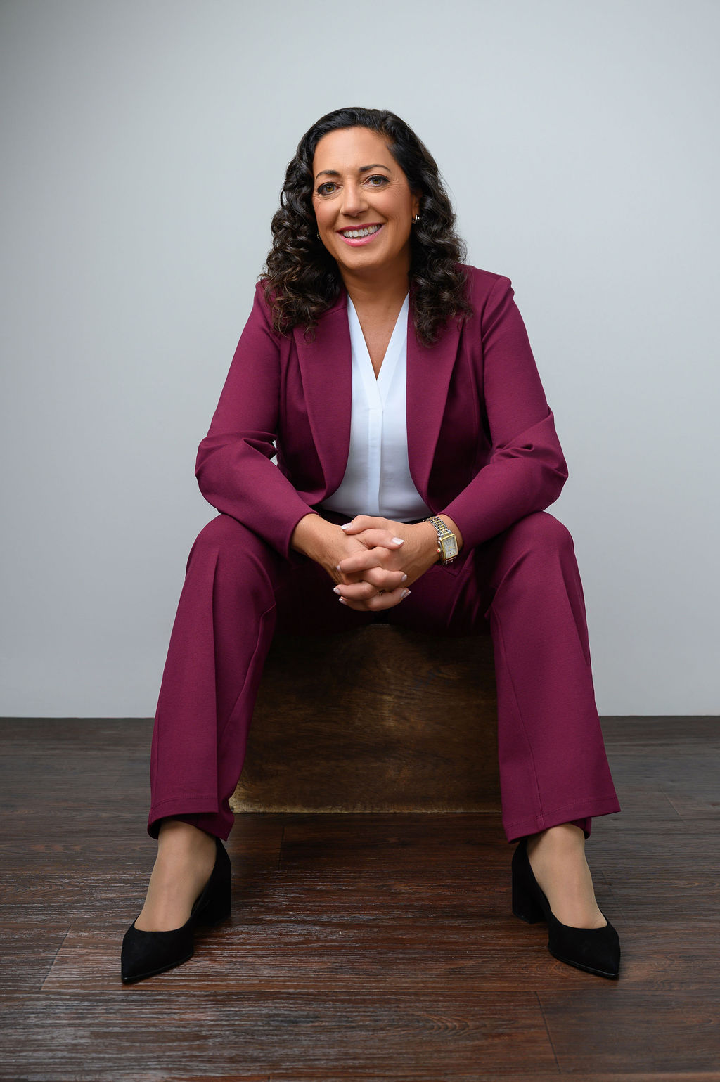 Seated professional portrait of a senior executive woman in a burgundy blazer smiling against a white background, photographed in New Jersey